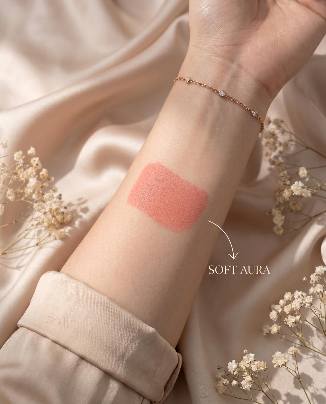 Hand with a swatch of pink lipstick labeled 'Soft Aura' on a soft background with flowers.