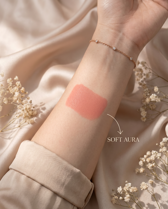 Hand with a swatch of pink lipstick labeled 'Soft Aura' on a soft background with flowers.