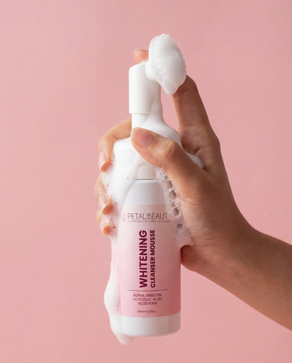 Hand holding a foamy bottle of Petal Beauty Whitening Cleanser Mousse against a pink background