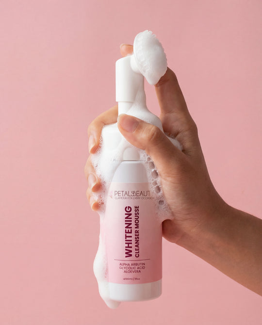 Hand holding a foamy bottle of Petal Beauty Whitening Cleanser Mousse against a pink background