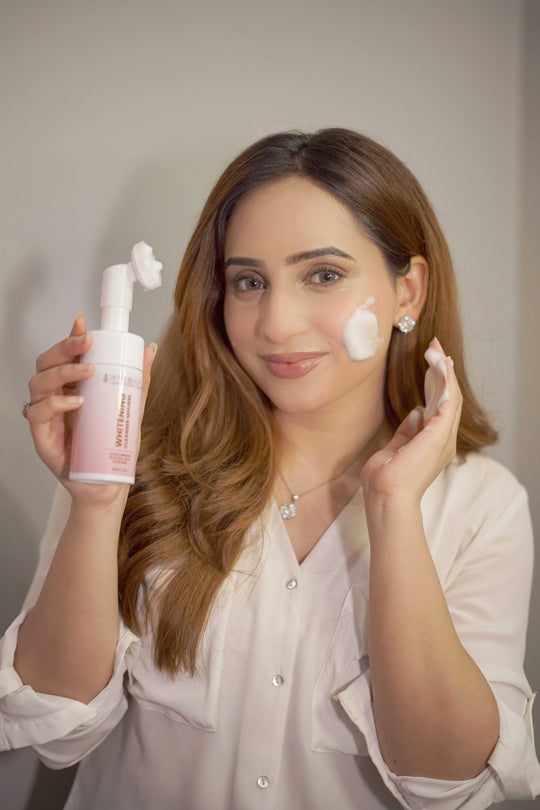 Woman applying a skincare product creating foam to her face with a bottle in hand against a neutral background
